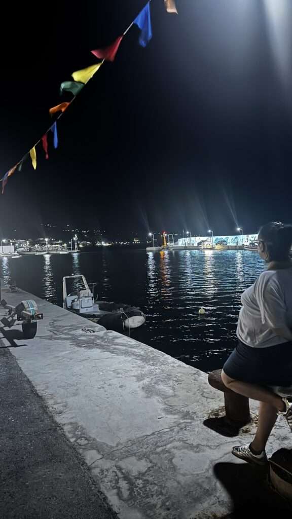 Maria assise au bord du port la nuit, sous des guirlandes de fanions colorés, en Crète