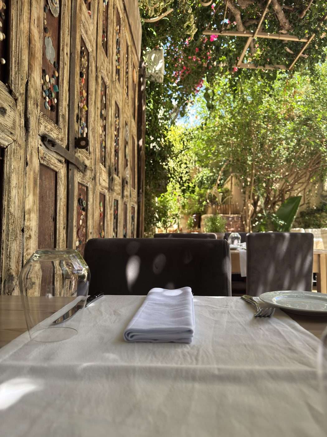 Table dressée avec nappe blanche dans le jardin du restaurant Avli à Réthymnon, vieilles portes en bois sculpté et treille de bougainvillier