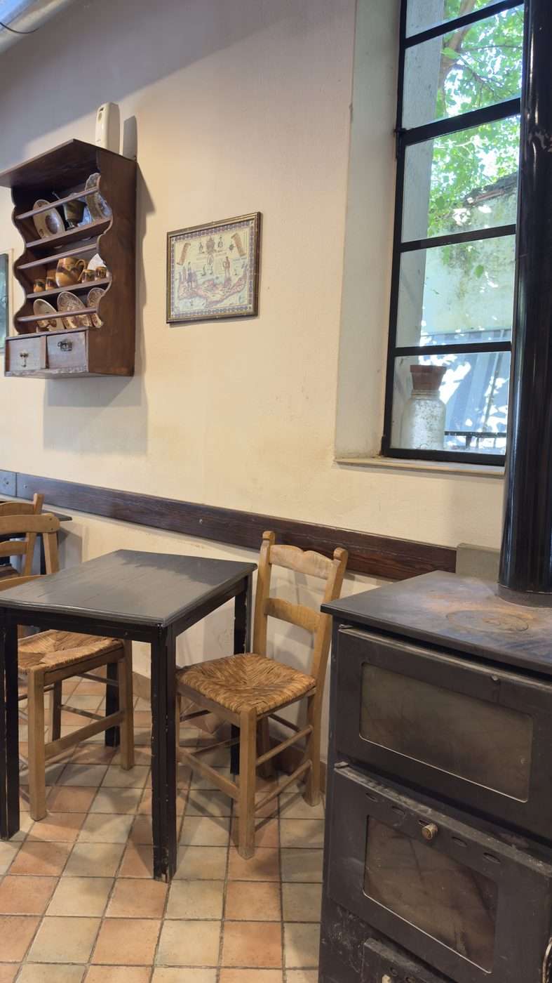 Intérieur d'un kafeneio crétois avec poêle à bois, tables et chaises paillées, étagère de vaisselle ancienne et carte de Crète encadrée au mur