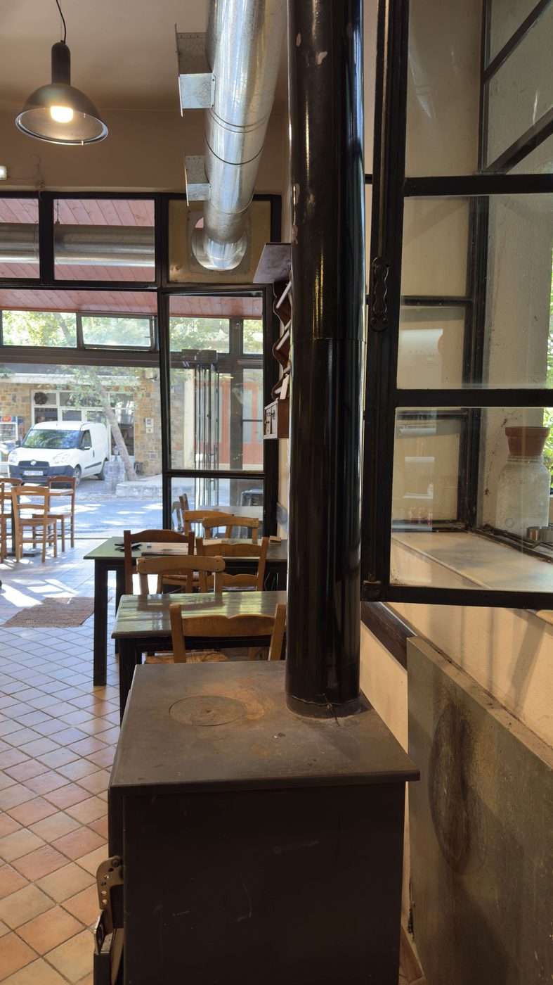 Salle d'un kafeneio crétois avec le poêle à bois au premier plan et la porte grande ouverte sur la rue du village