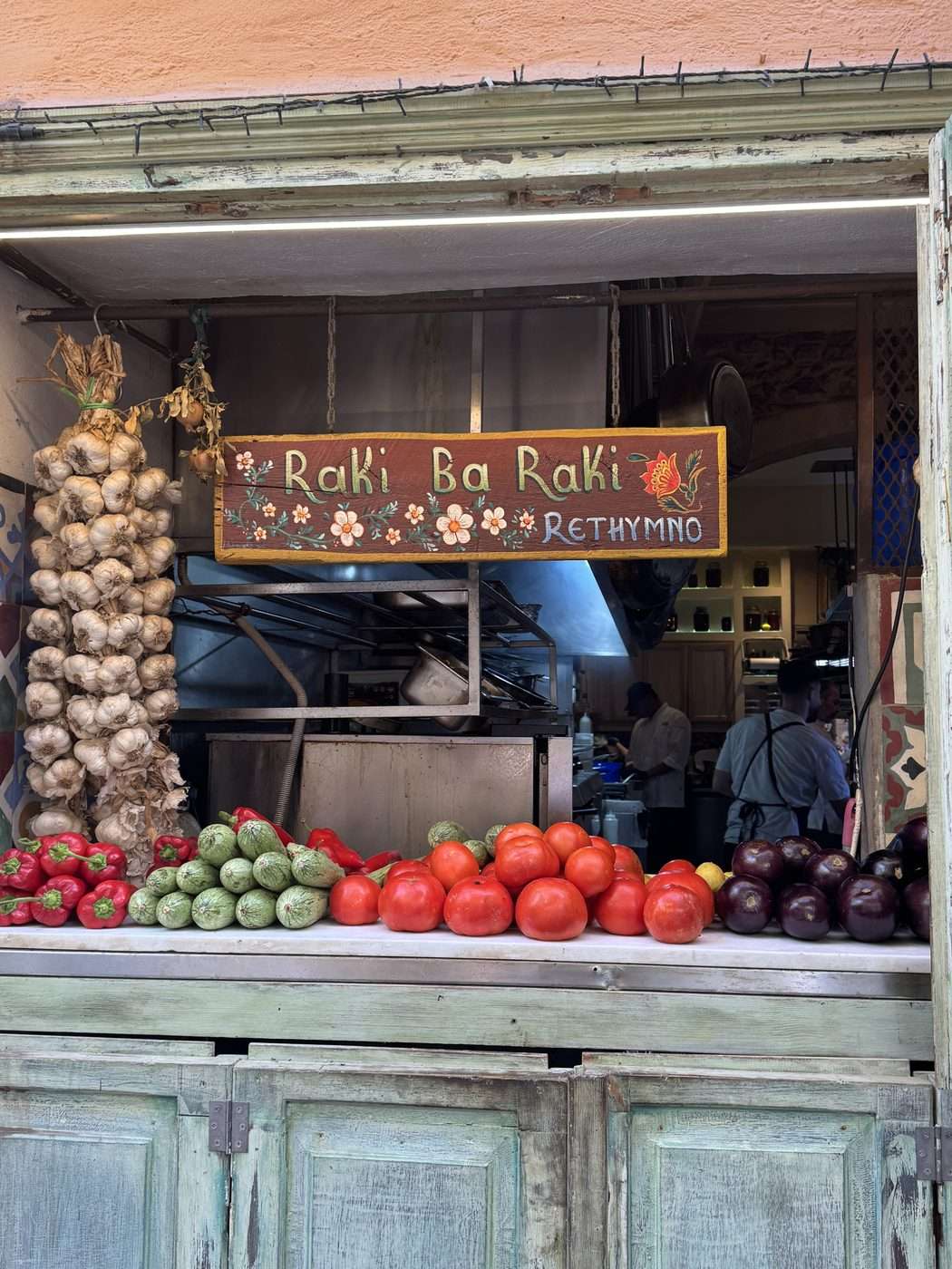 Entrée de la taverne Raki Ba Raki à Réthymnon en Crète, enseigne peinte à la main et guirlandes de tomates, aubergines, courgettes, poivrons et tresses d'ail devant la cuisine ouverte