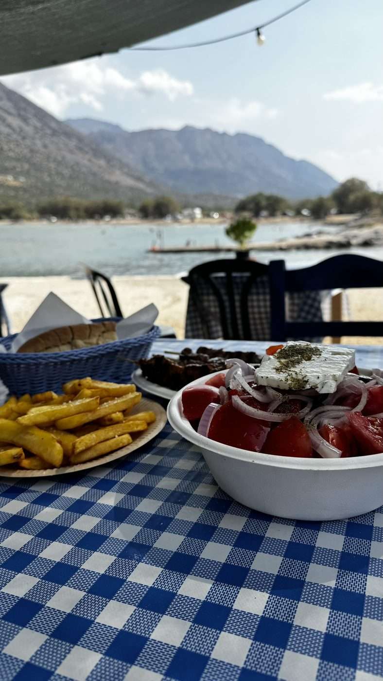 Table de taverne au bord de la mer en Crète, nappe vichy bleue, salade de tomates et feta, frites et pain, vue sur la plage et la montagne
