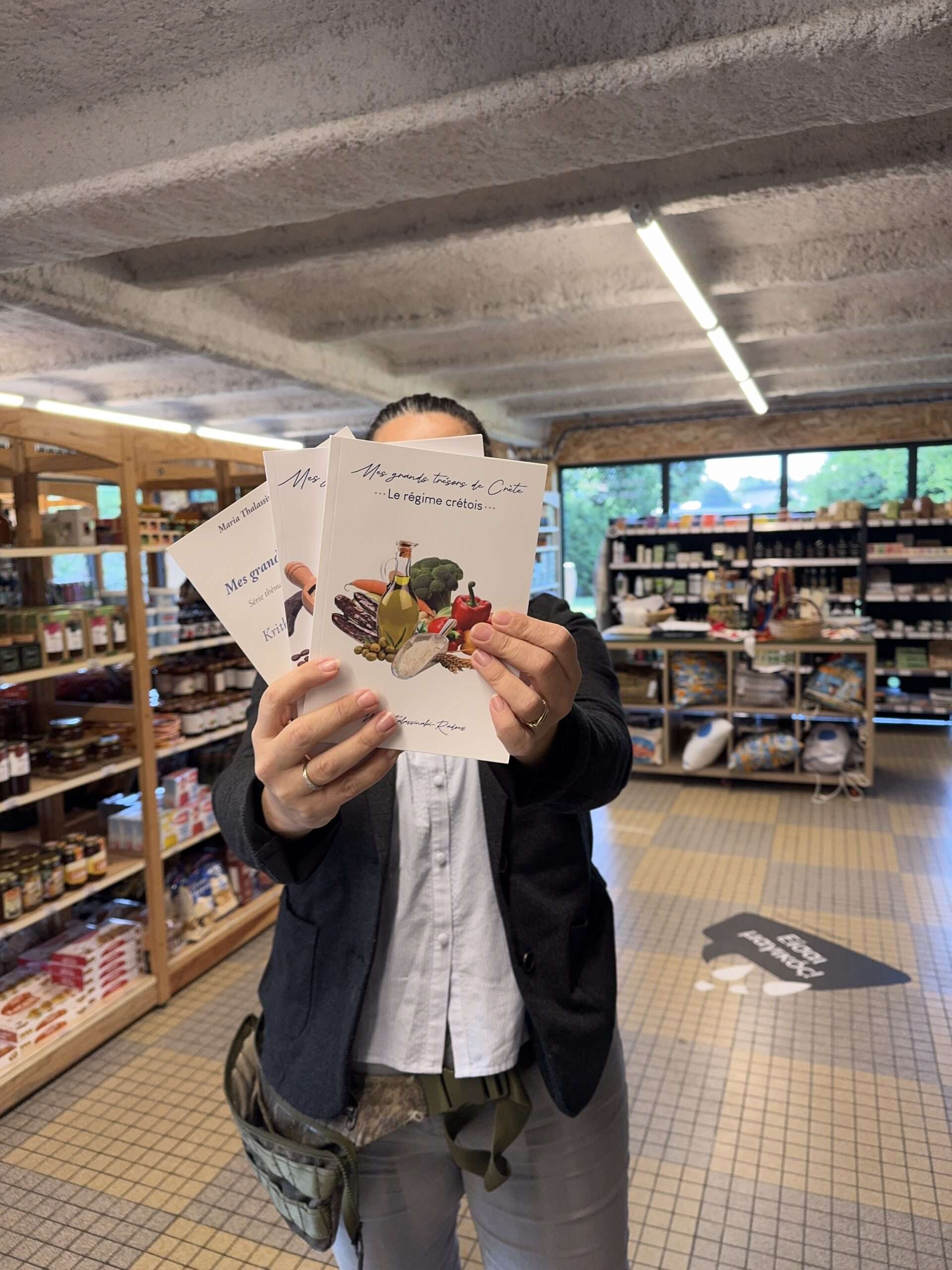 L'histoire de mes livres, du home déco au livre de cuisine grecque vegan 5 Maria cache son visage derrière ses trois livres, dans sa boutique de Messei