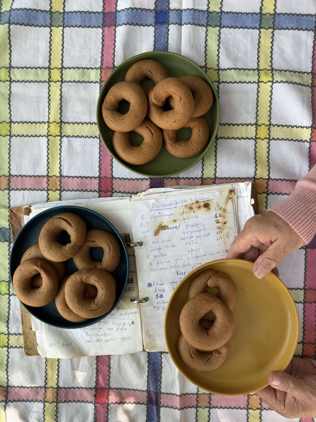 Grèce vegan, mon livre : la visite en avant-première, trois jours avant sa sortie 21 Des biscuits grecs en couronne sur des assiettes de couleur et un vieux cahier de recettes, une main tient l'assiette